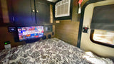 RV rental interior features a comfy bed with patterned linens, a mounted TV, overhead cabinets, AC unit, and a side door with a window.