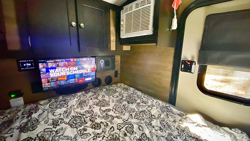 RV rental interior features a comfy bed with patterned linens, a mounted TV, overhead cabinets, AC unit, and a side door with a window. — photo 9
