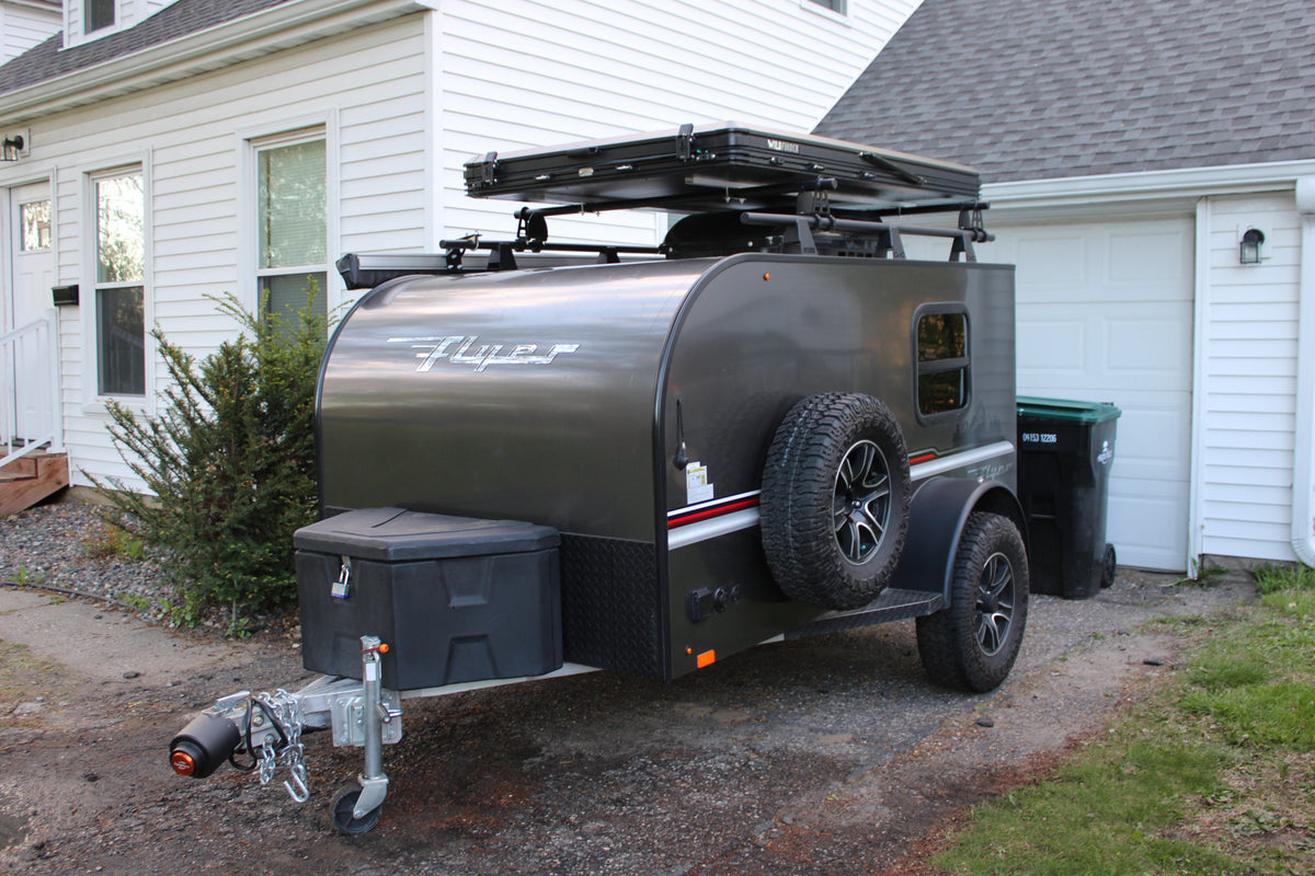 A dark gray Flyer camping trailer, equipped with a rooftop tent, is ready for rv rental adventures, parked on a gravel drive. — photo 6