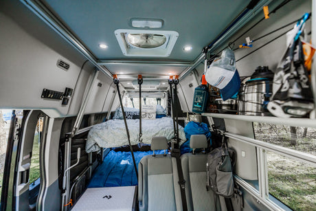 Interior view of a modern rv rental camper van. Features a raised bed in back, front seating, and abundant storage racks and shelves.