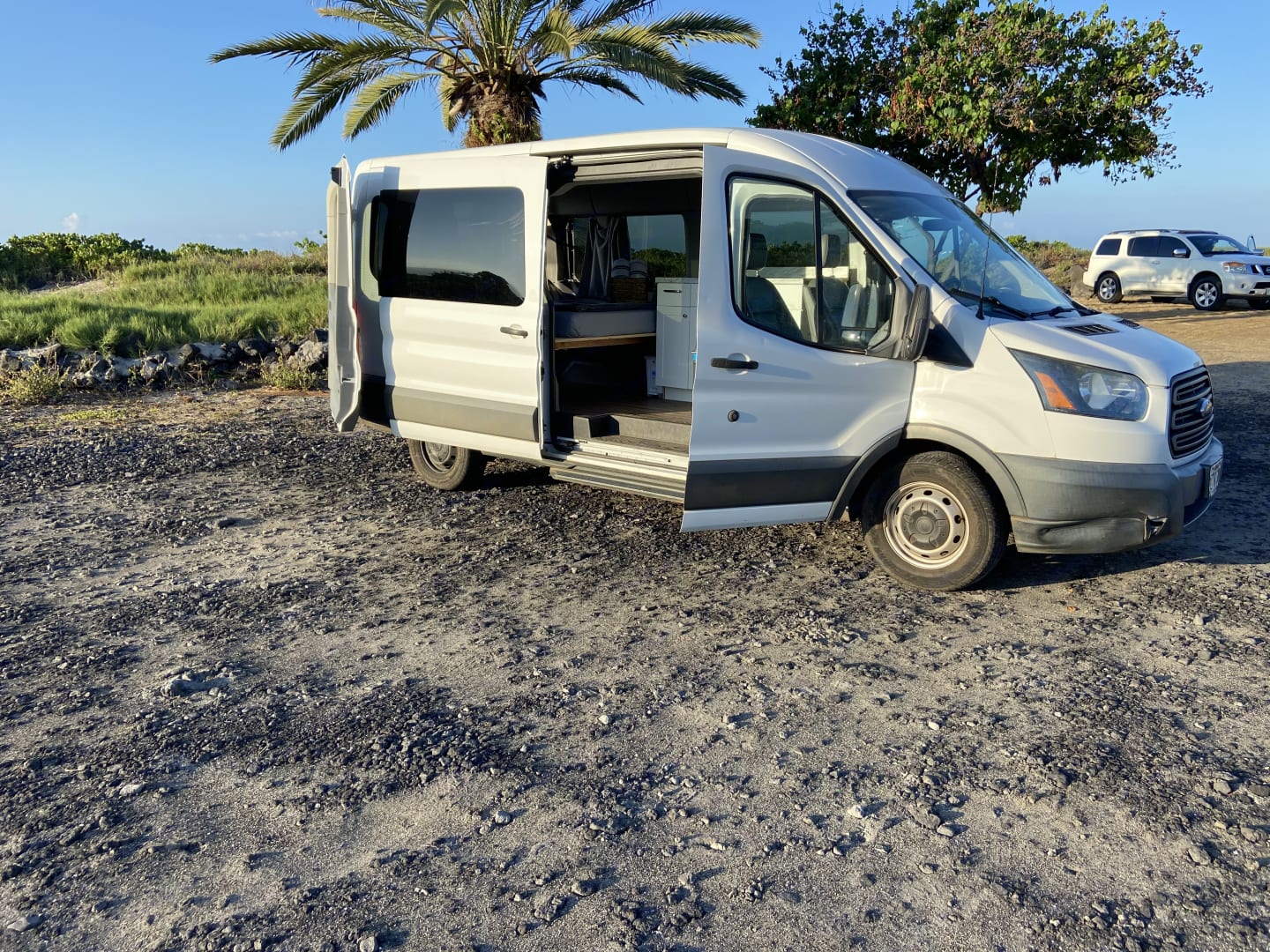 White Ford Transit camper van RV rental, side door open to reveal its custom interior, parked on a gravel lot near a palm tree.