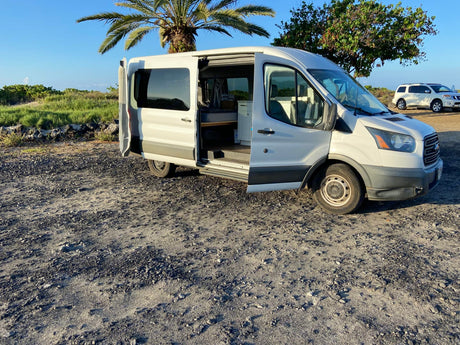 White Ford Transit camper van RV rental, side door open to reveal its custom interior, parked on a gravel lot near a palm tree.