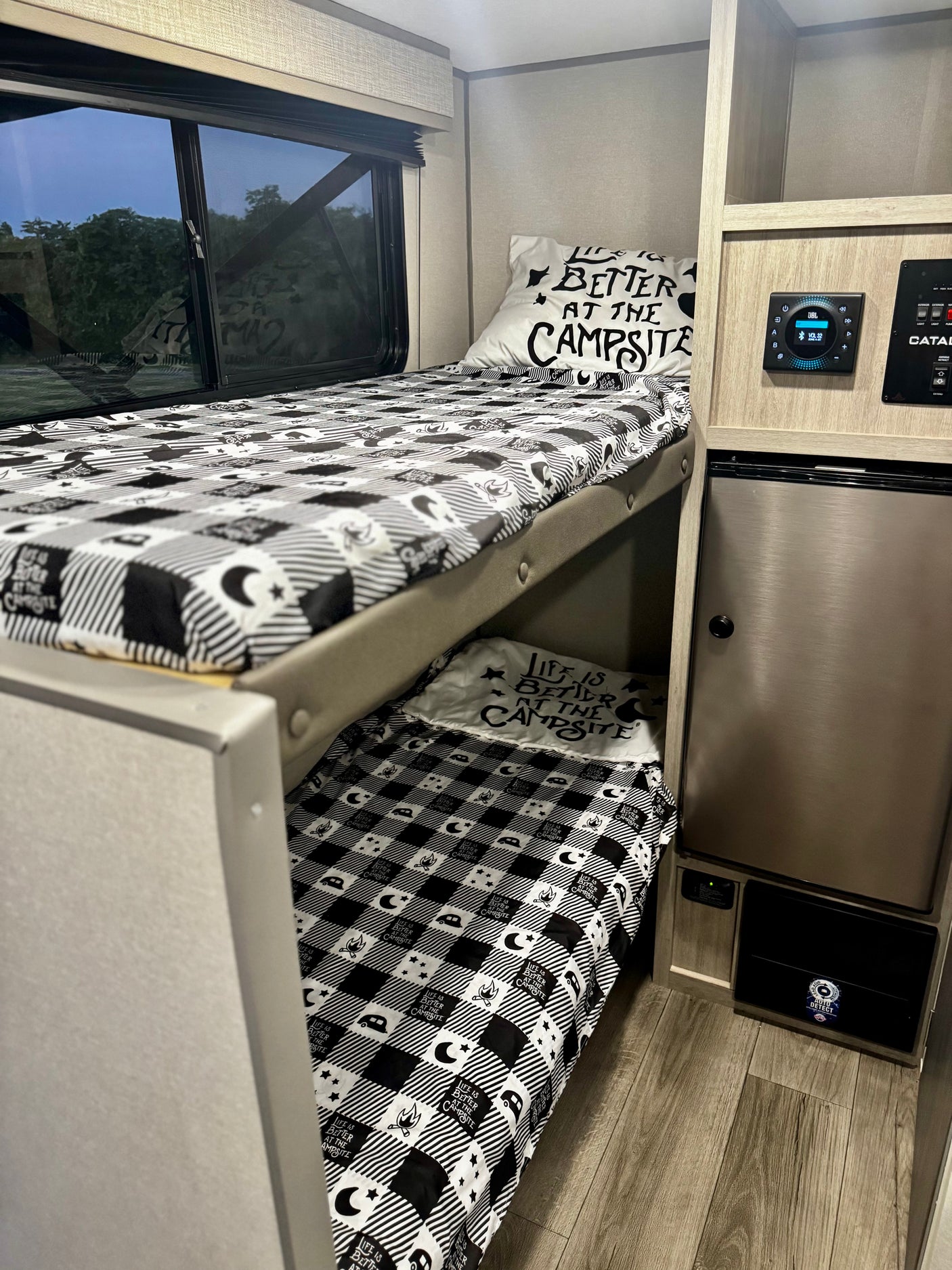 Inside an RV rental: two comfy bunk beds with black & white camping-themed bedding. Features include a window, JBL control, & fridge.