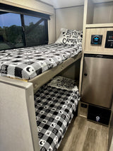 Inside an RV rental: two comfy bunk beds with black & white camping-themed bedding. Features include a window, JBL control, & fridge.