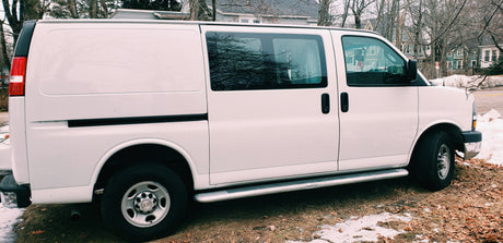 Side view of a white passenger van with tinted windows, parked on ground with leaves & snow. Consider this RV rental.