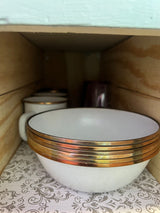 RV rental kitchen: Stacked white bowls with gold rims, plus mugs, in a wooden cabinet on a patterned liner.
