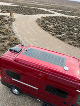 Overhead view of a red RV rental with solar panels on a remote gravel road, surrounded by dry, scrubby terrain.