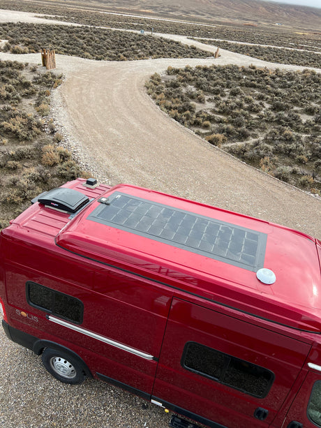Overhead view of a red RV rental with solar panels on a remote gravel road, surrounded by dry, scrubby terrain.