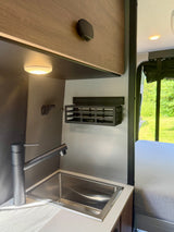 Modern RV rental kitchen with sink, black faucet, overhead storage. Green views & bed visible on the side.