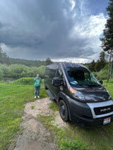 Stormy Idaho skies over an RV rental van parked by a river & forest, with a smiling woman in a green hoodie beside it.