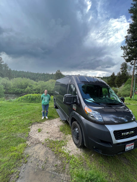 Stormy Idaho skies over an RV rental van parked by a river & forest, with a smiling woman in a green hoodie beside it.