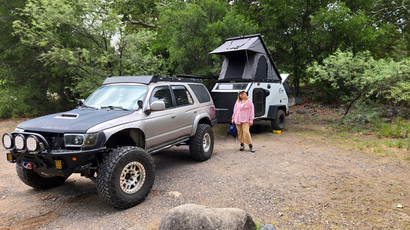 Off-road RV rental setup: a modified 4Runner and XTR camper with a rooftop tent at a wooded campsite, with a woman standing by. — photo 14