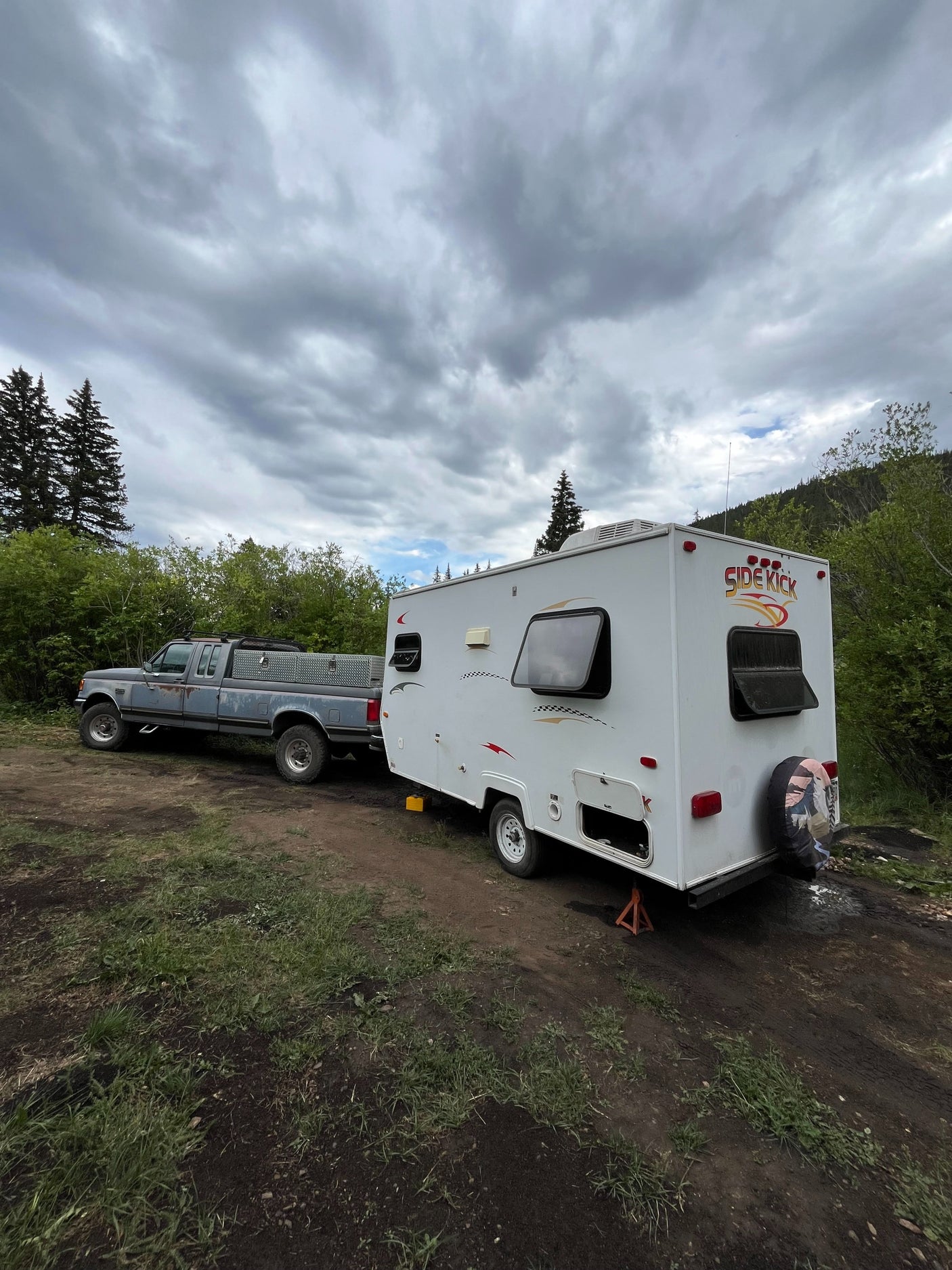 RV rental: A truck tows a Side Kick travel trailer on a dirt road, ready for camping under a cloudy sky.