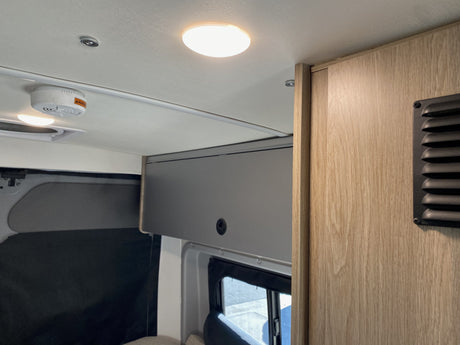 Brightly lit RV rental interior showing a ceiling light, smoke detector, gray overhead cabinet, and wood-grain wall.