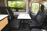 RV rental interior: dining area with white table, black bench seat, and swivel seat. Window view of trees.