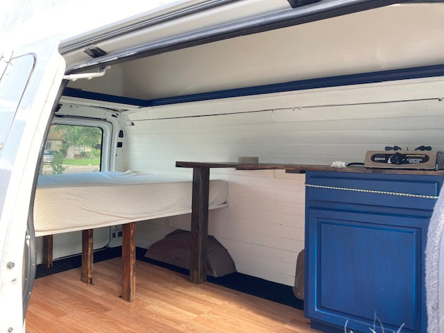 RV rental van conversion interior featuring a cozy bed, wood floor, and blue kitchen cabinet with camping stove.