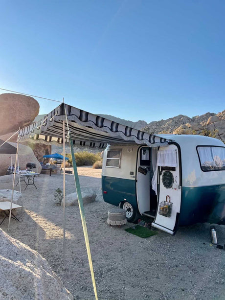 Teal & white rv rental with striped awning set up at a sunny, rocky desert campsite. Tables & tents nearby.