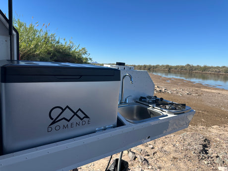 RV rental outdoor kitchen featuring a portable fridge, sink, and stove set by a calm river.