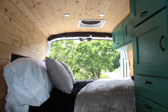 This RV rental interior features a cozy bed with pillows, wood-paneled walls/ceiling, green cabinets, and a serene outdoor view.