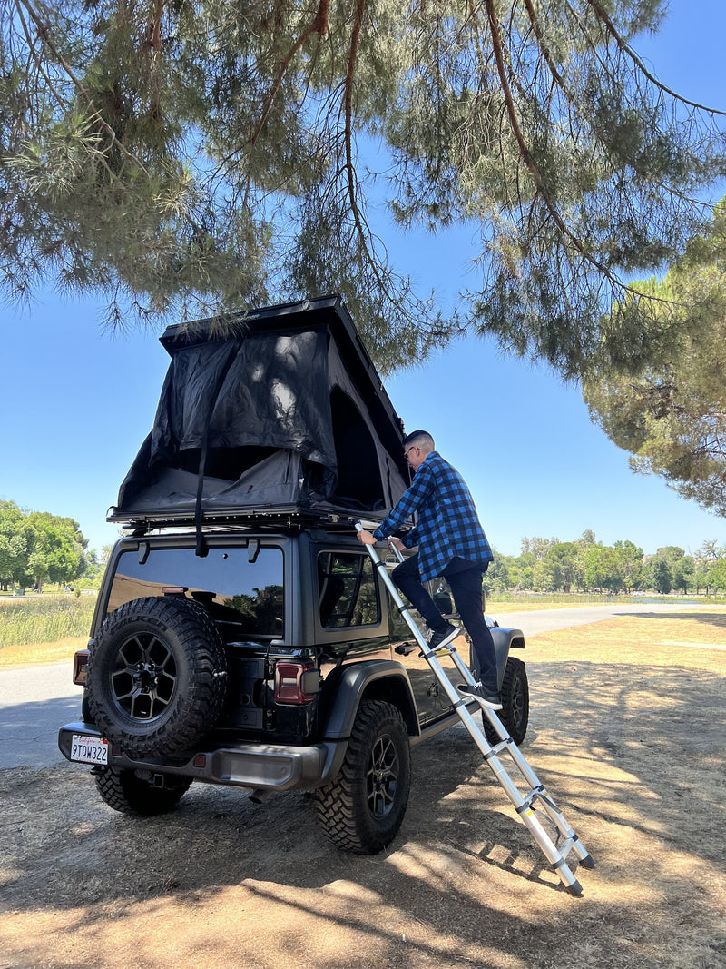 Setup in progress on a dirt pullout, the seller on the telescoping ladder securing the opened tent, showing the ladder angle and the tent's standing height above the vehicle — photo 5