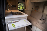 Compact RV rental interior showcasing a kitchen with a pull-out cutting board, stove, and a comfortable sofa with cup holders.