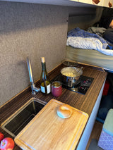 Cozy RV rental interior shows a compact kitchen with cooking pasta, sink, wine, and a bed in the background.
