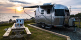 A sleek Airstream RV rental sits oceanside at sunset, awning extended, with a picnic table and cooler ready for camp.