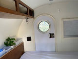 Bright rv rental interior. Features a unique porthole door, bed, wooden cabinets, and a potted plant on a built-in shelf.