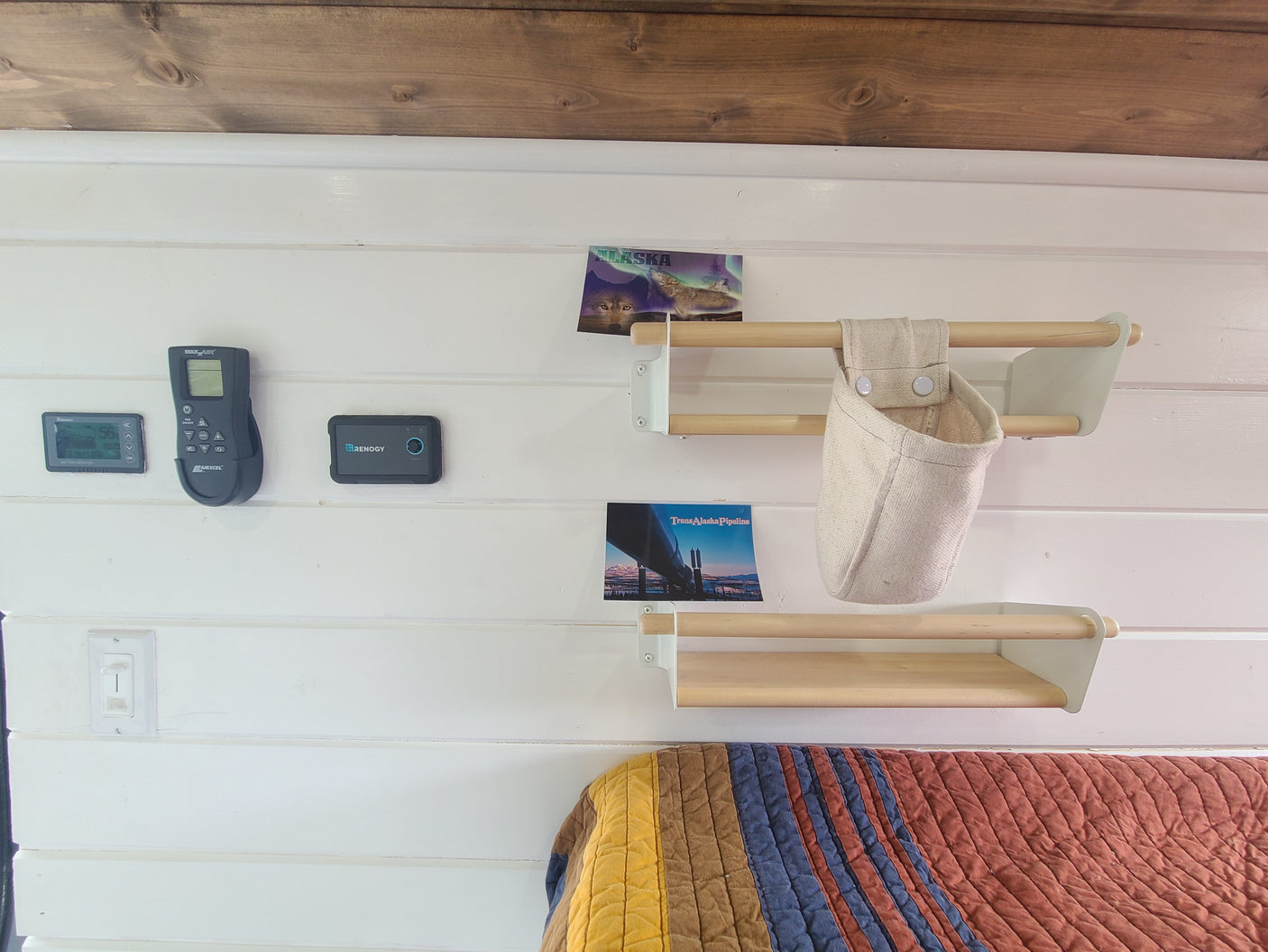 An RV rental's cozy interior featuring a fan remote, battery monitor, small shelves, travel postcards, and a quilted bed.