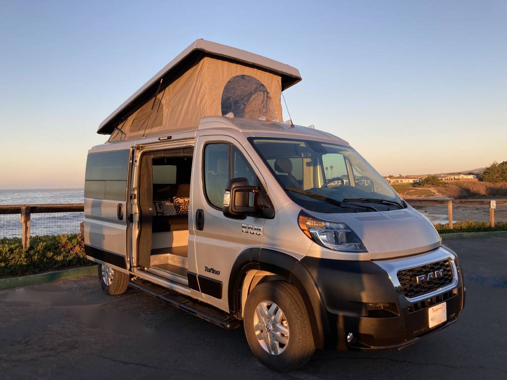 This silver pop-top camper van rv rental offers ocean views at sunset. Perfect for a coastal road trip adventure.|RV rental interior with stunning ocean views from pop-top clear and mesh windows. Includes a bed for coastal camping.|A silver pop-top RV rental van parked in a snow-dusted forest campsite next to a mountain. Ideal for winter adventures.|Rear view of an RV rental with two bikes secured on a hitch-mounted rack
