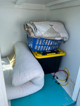 Organized RV rental storage shows rolled mattress, quilted blankets, a storage bin with tools, and bungee cords.