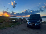 Experience a beautiful lake sunset with this RV rental. Black van parked on gravel, serene waters, vibrant sky.