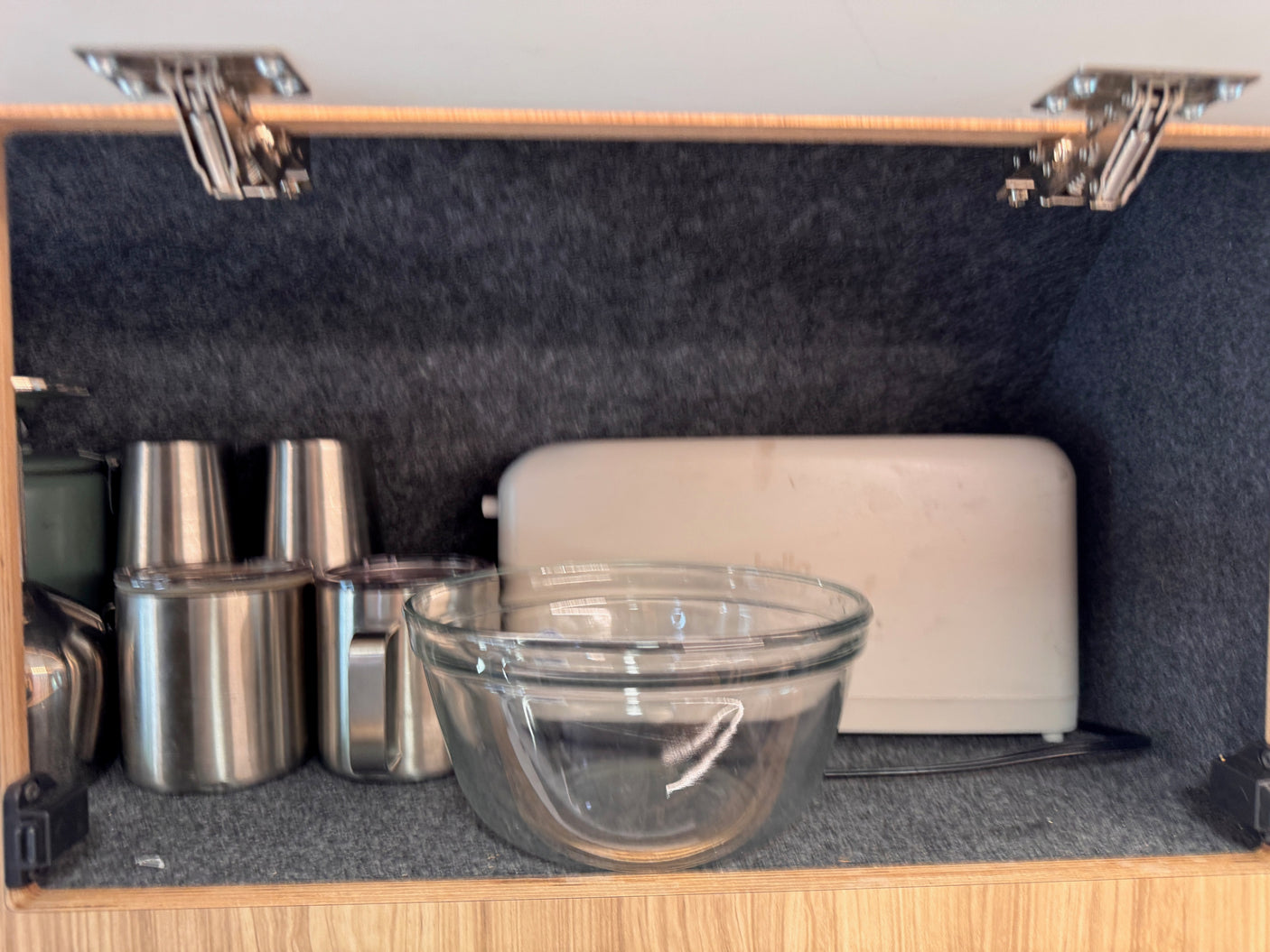 Inside an RV rental cabinet: stainless steel cups, a glass bowl, and a toaster on a felt-lined shelf.