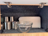 Inside an RV rental cabinet: stainless steel cups, a glass bowl, and a toaster on a felt-lined shelf.