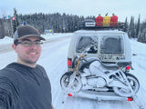Adventure awaits! A man smiles in front of a snow-covered van and motorcycle, likely an RV rental, at the Alaska border in winter.