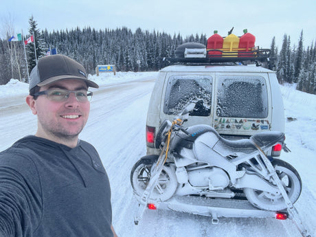 Adventure awaits! A man smiles in front of a snow-covered van and motorcycle, likely an RV rental, at the Alaska border in winter.