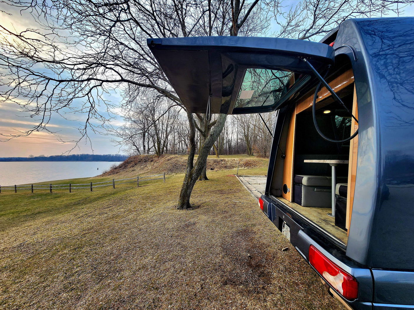 RV rental camper van parked by a serene lake at sunset, with its rear open revealing a cozy interior table setup.
