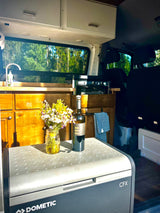 Inside an RV rental: a bright kitchen setup with a Dometic fridge, flowers, and a forest view.