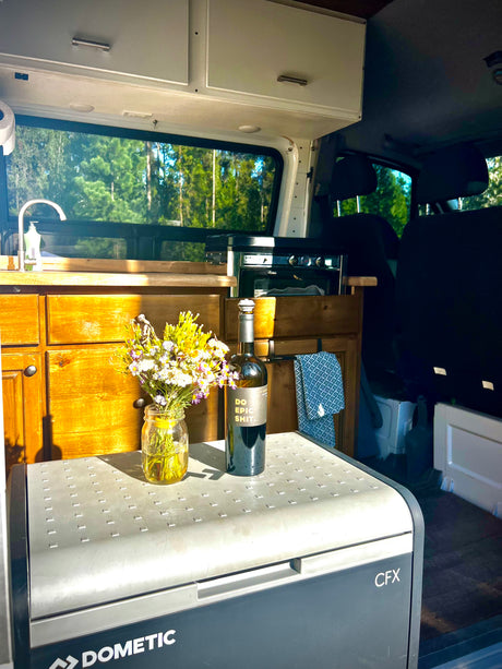 Inside an RV rental: a bright kitchen setup with a Dometic fridge, flowers, and a forest view.