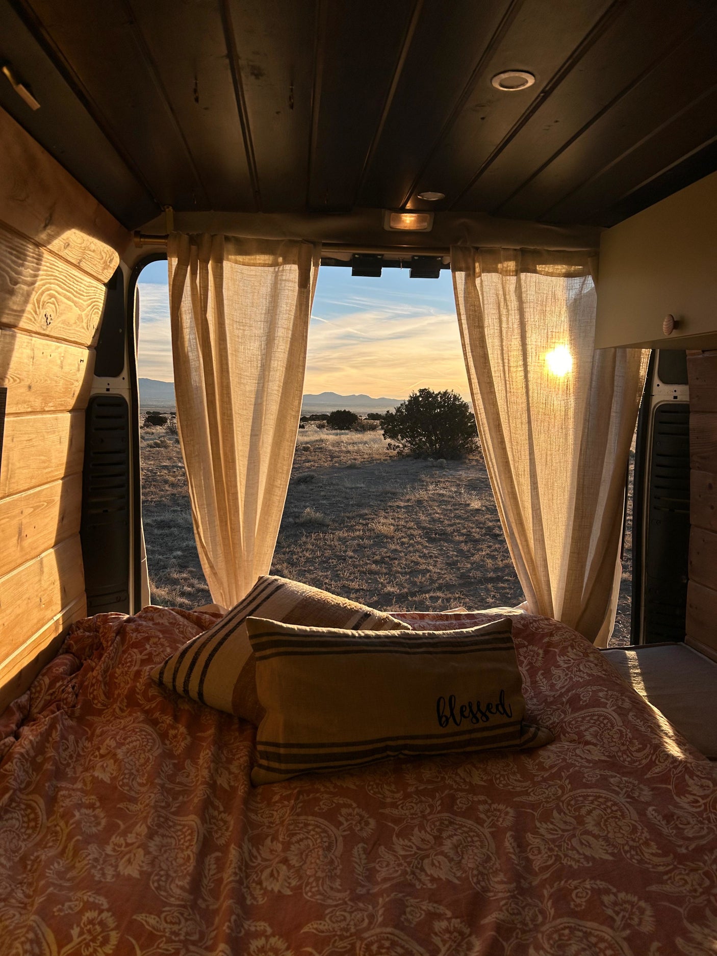 Cozy RV rental interior offers a stunning sunset view over a desert landscape through open back doors, framed by curtains and golden light.
