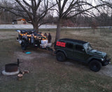 An overland Jeep tows a compact off-grid camper/rv rental, lit with string lights at a natural campsite with a fire pit.