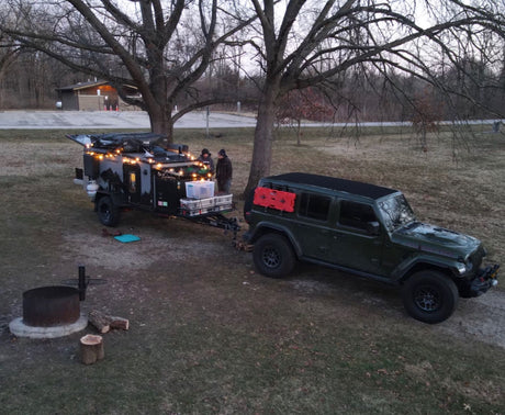 An overland Jeep tows a compact off-grid camper/rv rental, lit with string lights at a natural campsite with a fire pit.