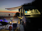 An RV rental campervan lights up a beach evening with a person, dog, and warm campfire at sunset.