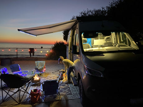 An RV rental campervan lights up a beach evening with a person, dog, and warm campfire at sunset.