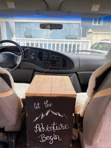 Interior view of an RV rental van, looking forward. Features front seats, dashboard, and a decorative "Let the Adventure Begin" sign.
