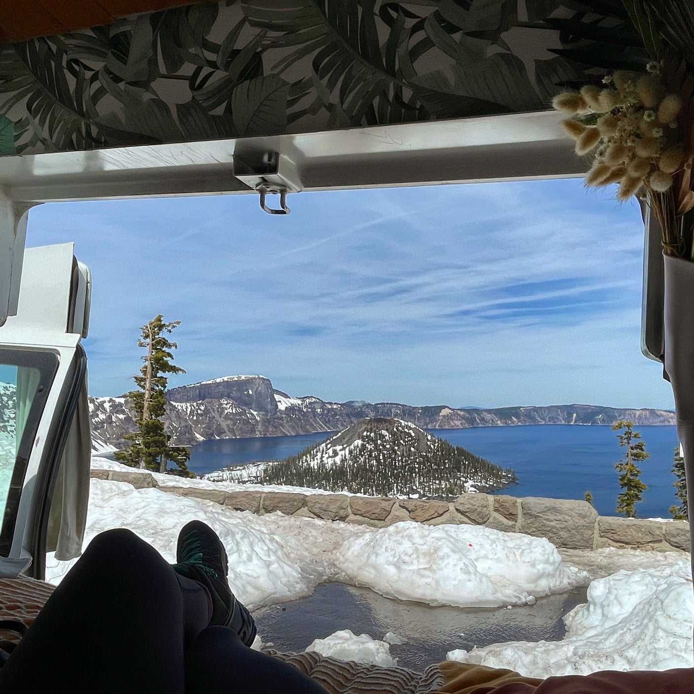 Person's legs propped up inside an RV rental overlooking snowy Crater Lake.