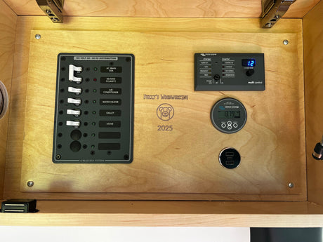 This RV rental's power hub features a Blue Sea AC panel, Victron inverter control, and battery monitor displaying voltage, all on wood.