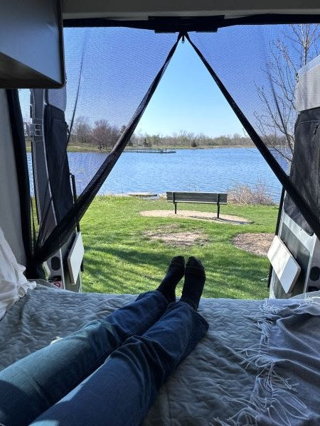 RV rental comfort: Legs on bed inside, looking out through screened doors at a calm lake, distant pier, and park bench.