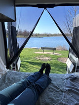 RV rental comfort: Legs on bed inside, looking out through screened doors at a calm lake, distant pier, and park bench.
