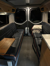 RV rental interior: dark benches with wood table, textured floor, rear windows. Overhead storage completes this mobile space.